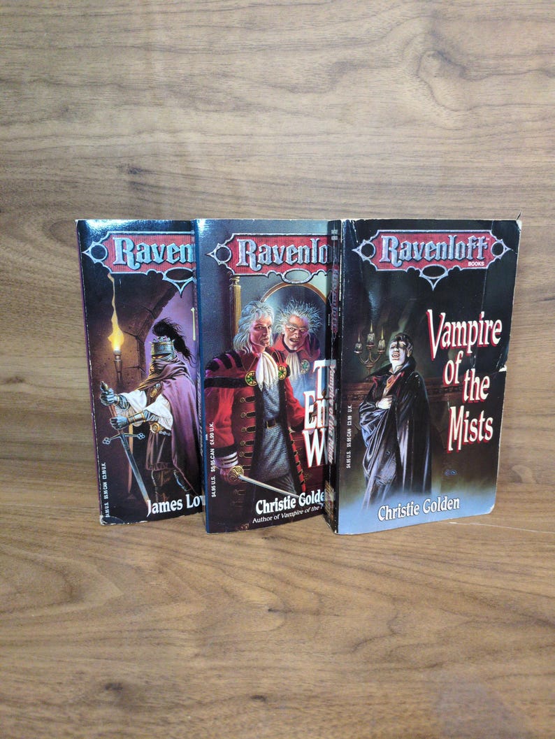 Puede incluir: Se muestran tres libros de Ravenloft, con ilustraciones g&oacute;ticas y t&iacute;tulos como "Vampire of the Mists". Las portadas presentan im&aacute;genes oscuras con figuras y texto, sobre un fondo de madera. Los autores son James Lowder y Christie Golden.