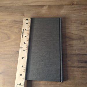 May include: A gray book with a textured cover is next to a wooden ruler. The ruler is marked with inches, showing the book is approximately 9 inches long. The book is on a wooden surface.
