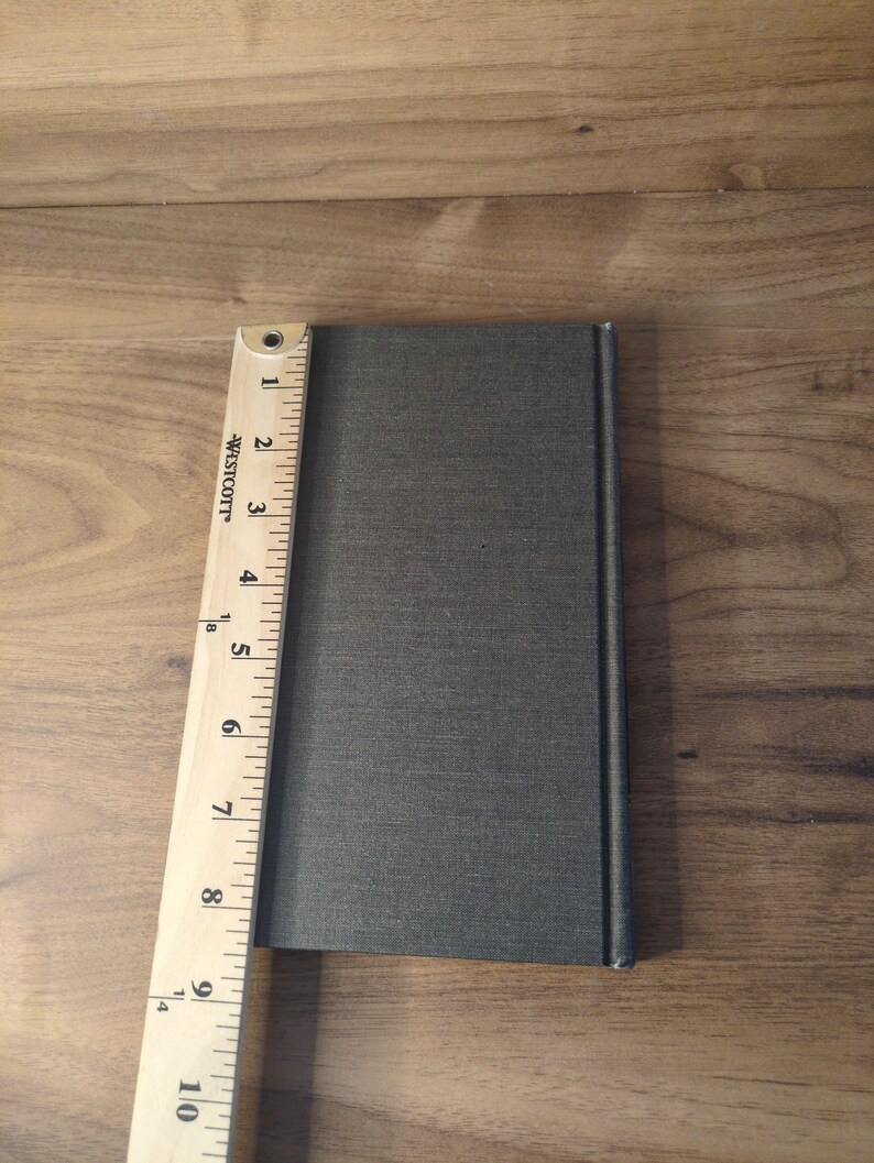 May include: A gray book with a textured cover is next to a wooden ruler. The ruler is marked with inches, showing the book is approximately 9 inches long. The book is on a wooden surface.