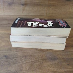 Puede incluir: Tres libros de bolsillo apilados sobre una superficie de madera. El libro superior tiene una cubierta oscura con el t&iacute;tulo "Knight of the Black Rose" visible. El lomo del libro es visible y las p&aacute;ginas son de color blanquecino. Los libros est&aacute;n en buen estado.