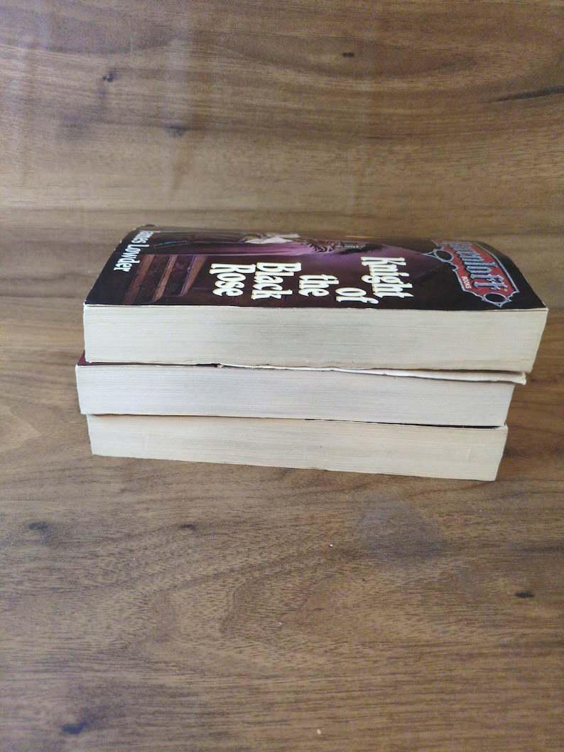 Puede incluir: Tres libros de bolsillo apilados sobre una superficie de madera. El libro superior tiene una cubierta oscura con el t&iacute;tulo "Knight of the Black Rose" visible. El lomo del libro es visible y las p&aacute;ginas son de color blanquecino. Los libros est&aacute;n en buen estado.
