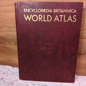 May include: A maroon-colored "Encyclopædia Britannica World Atlas" book. The book's cover features gold lettering and a textured surface. The book is resting on a white lace tablecloth, with a wooden background.