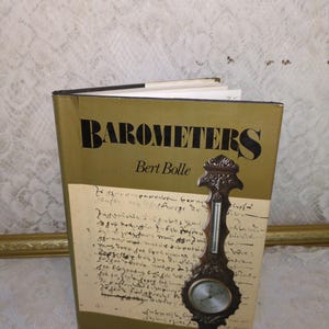 May include: A vintage book titled "Barometers" by Bert Bolle, with a decorative, ornate barometer attached to the cover. The book has a gold cover with black lettering and a cream-colored page with handwritten script.