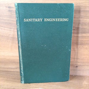May include: A hardcover book titled "SANITARY ENGINEERING" in white lettering on a dark teal cover. The book is standing upright on a wooden surface. The book's spine is visible, showing the binding.