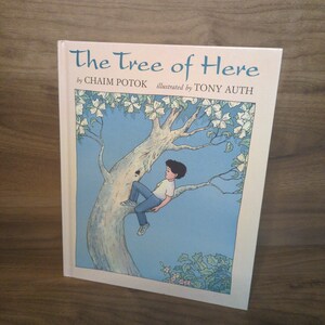 May include: A hardcover book titled The Tree of Here by Chaim Potok and illustrated by Tony Auth. The cover features a drawing of a person with dark hair wearing a white shirt and blue jeans sitting in a large tree with white flowers against a blue sky.