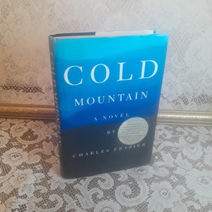 May include: A hardcover book titled "Cold Mountain" by Charles Frazier. The cover features a blue and black gradient design with the title in white text.