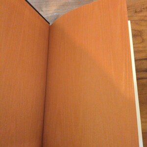 May include: An open book with orange-colored pages and a black spine. The pages have a textured appearance, and the book is positioned on a wooden surface. The book's pages are a warm, inviting color.