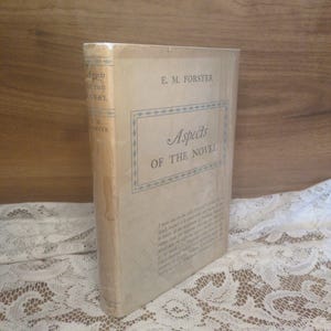 May include: A white ceramic mug with a black handle and the text "But First, Coffee" in a bold, black font. The mug is set against a blurred background, suggesting a cozy setting.