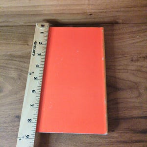 May include: An orange rectangular object, approximately 25 cm long, is positioned next to a wooden ruler. The object has a smooth, solid color and appears to be a book or similar item. The ruler is marked with inch measurements.