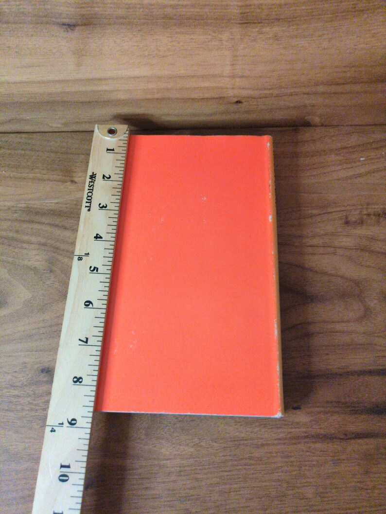 May include: An orange rectangular object, approximately 25 cm long, is positioned next to a wooden ruler. The object has a smooth, solid color and appears to be a book or similar item. The ruler is marked with inch measurements.