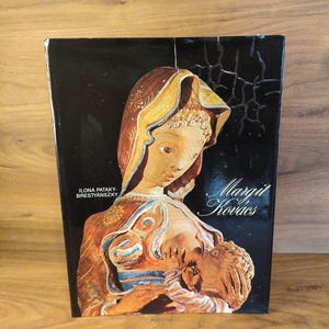 May include: A book with a black cover featuring a relief sculpture of a woman holding a child. The sculpture is in shades of brown, beige, and gold. The book's title is "Margit Kovacs."
