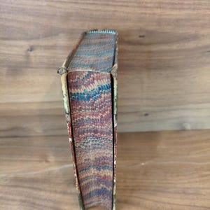 May include: A vintage book with a marbled page edge design featuring swirling patterns of red, blue, and cream colors. The book spine is bound in a light-colored material with visible wear along the edges and corners of the cover structure.