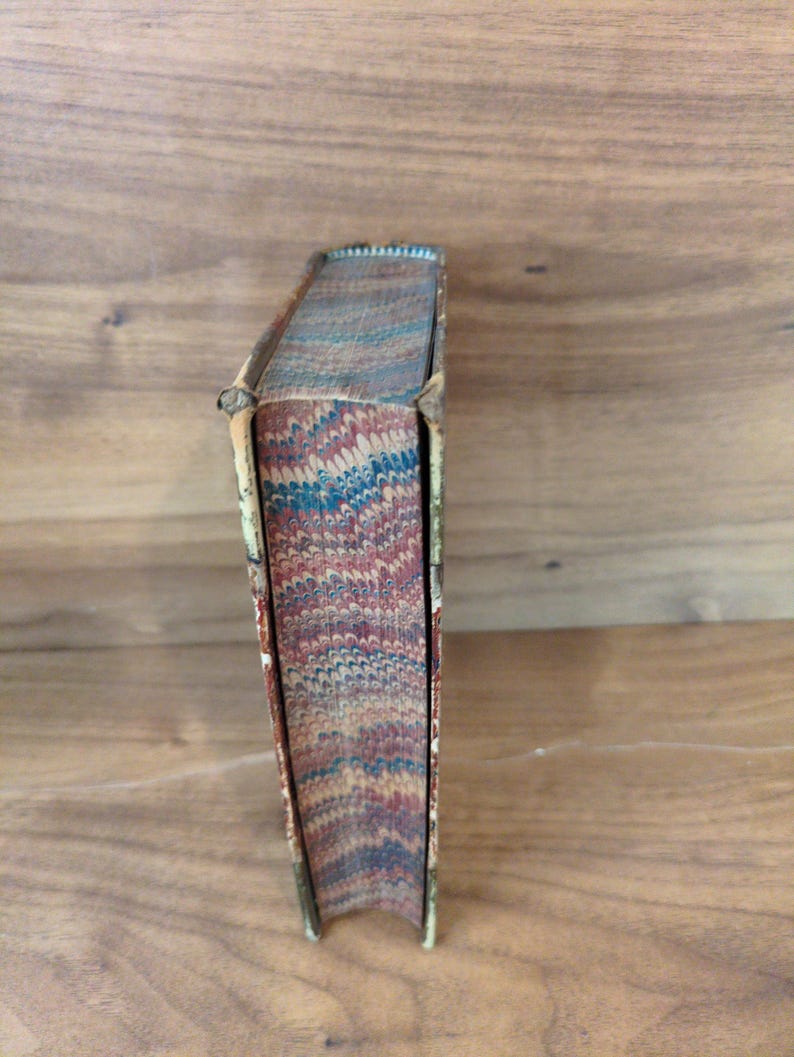 May include: A vintage book with a marbled page edge design featuring swirling patterns of red, blue, and cream colors. The book spine is bound in a light-colored material with visible wear along the edges and corners of the cover structure.