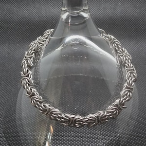 May include: A silver-toned chainmail bracelet featuring a complex Byzantine weave pattern. The bracelet is constructed from numerous small, interlocking metal rings that create a continuous, textured, and flexible circular band design.