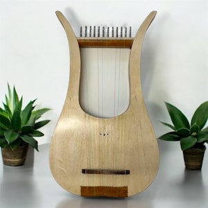 May include: A light wood lyre with 15 strings, a wooden soundboard, and a brown wooden bridge. The lyre is sitting on a table with two green plants in pots.