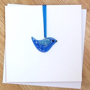 Puede incluir: Adorno brillante en forma de pájaro azul, con un diseño punteado en el pecho, suspendido de una cinta azul a juego. El pájaro se encuentra frente a tarjetas blancas, lo que sugiere una tarjeta de felicitación o una etiqueta de regalo. Diseño sencillo.