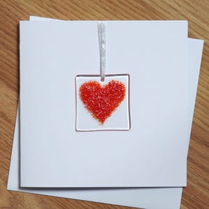 Puede incluir: Adorno cuadrado de vidrio transparente con un diseño de corazón rojo. El corazón está lleno de pequeñas partículas rojas texturizadas. Se adjunta una cinta blanca para colgarlo. El adorno se muestra en una tarjeta blanca.