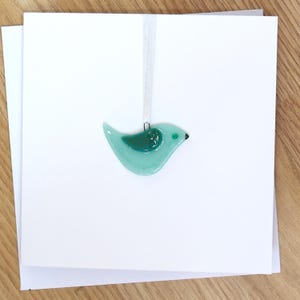 Puede incluir: Adorno de pájaro de cristal turquesa con una cinta blanca, colgado frente a una pila de tarjetas blancas. El pájaro tiene un patrón verde oscuro en su ala y un pico negro. El fondo es una superficie de madera clara.
