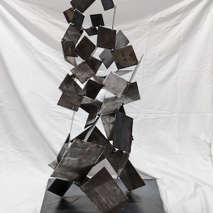 May include: A modern metal sculpture made of many rectangular pieces of metal welded together. The sculpture is abstract and has a dark gray finish.