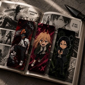 Könnte beinhalten: Drei rechteckige Lesezeichen mit Anime-Figuren auf einem aufgeschlagenen Manga-Buch. Das erste Lesezeichen zeigt eine Figur mit Kettensägenkopf, das zweite eine Figur mit langem blonden Haar und Hörnern, das dritte eine Figur, die eine Waffe hält.