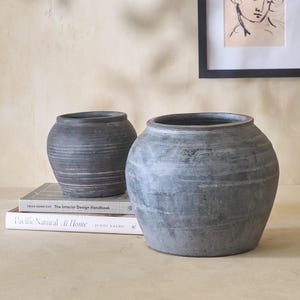 May include: Two gray ceramic pots, one slightly larger than the other, with a rough, textured surface. The pots are sitting on a table with two books stacked on top of each other.