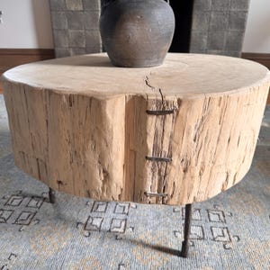 May include: A rustic, round coffee table made from a section of a tree trunk. The light-colored wood has visible grain and is secured with metal staples. It rests on three dark metal legs, with a dark vase on top.