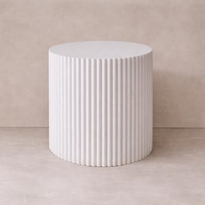 May include: A white, cylindrical side table with a fluted design. The table has a round top and a series of vertical, ridged lines that run down the sides. The table is approximately 20 inches tall and 18 inches in diameter.