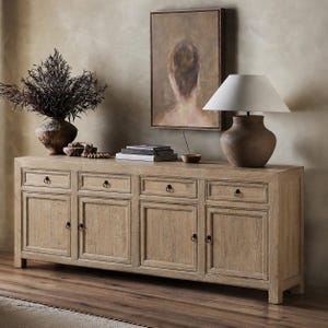 May include: A long, light-toned wooden sideboard featuring four drawers and four cabinet doors, each equipped with small, dark circular metal pulls. The piece has a rectangular frame with visible wood grain and sits on short, sturdy legs.