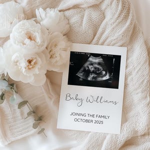 May include: A white card with a black and white ultrasound image and the text "Baby Williams Joining the Family October 2025". The card is on a white knitted blanket with white flowers.