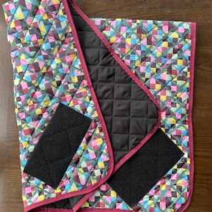 Peut inclure: Coussinet matelassé avec un motif géométrique dans les tons de rose, bleu, jaune et vert. Le coussinet a un intérieur noir et est bordé d'une bordure bordeaux. Deux sangles velcro noires sont attachées.