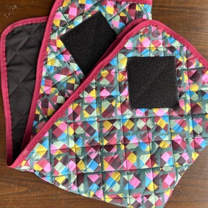 Peut inclure: Deux tapis de selle matelassés en tissu avec un motif géométrique dans des tons de rose, bleu, jaune et vert. Les tapis ont une bande de velcro noire de chaque côté.