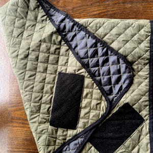 May include: Olive green quilted fabric with a diamond pattern. The edges are trimmed with black fabric, and there are two black rectangular patches. The fabric is folded over, revealing a black quilted lining.