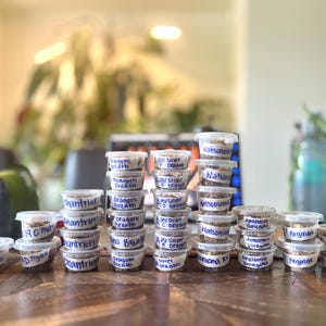 Peut inclure: Plusieurs petits récipients en plastique transparent remplis d'une substance brune, empilés sur une table en bois. Chaque récipient porte une étiquette manuscrite avec des noms tels que "Dragons Breath" et "Watsoniana".