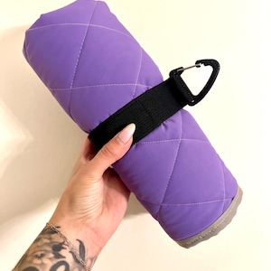 Peut inclure: Une couverture matelassée violette enroulée avec un motif de surpiqûres en losange. La couverture est maintenue par une sangle en nylon noir dotée d'un mousqueton en plastique noir fixé sur le côté pour faciliter le transport et le rangement.