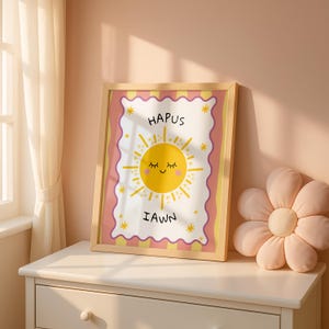 Puede incluir: Una impresión artística enmarcada con un sol amarillo sonriente y las palabras "Hapus Iawn". La impresión tiene un borde rosa y amarillo y se exhibe en una cómoda blanca. Una almohada rosa con forma de flor está al lado del marco.