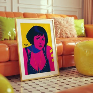 May include: A framed art print featuring a pop art style portrait of a woman with dark hair and a blue and pink color scheme. The artwork is set against a yellow background and is in a gold frame. The word "TIDY" is in the corner.