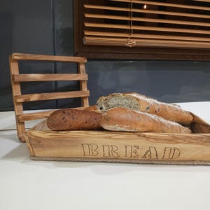 以下が含まれることがあります： 自然な木目模様の長方形の木製トレイで、前面にBREADという文字が焼き付けられています。トレイには垂直な木製の格子状の背もたれがあり、中には暗い斑点が見えるカリッとしたパンが3つ入っています。