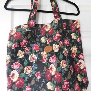 May include: A black tote bag featuring a floral pattern with red, pink, and cream-coloured roses, green leaves, and small blue berries. The bag has two fabric handles and a circular brown leather-like patch centred on the front of the exterior.