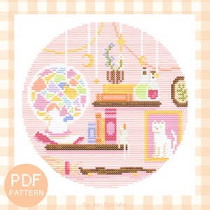 Peut inclure: Un modèle de point de croix représentant une pièce rose avec une étagère, un chat, une plante en pot et une lampe en verre coloré. Le motif est de forme circulaire et a un fond rose.
