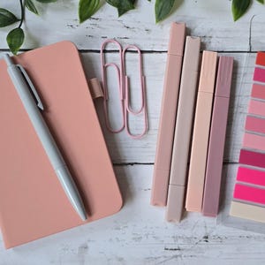 May include: A peach-coloured notebook with a grey pen, two light pink paper clips, four rectangular markers in shades of peach and mauve, and a clear plastic strip containing ten adhesive page markers in various shades of pink and red.