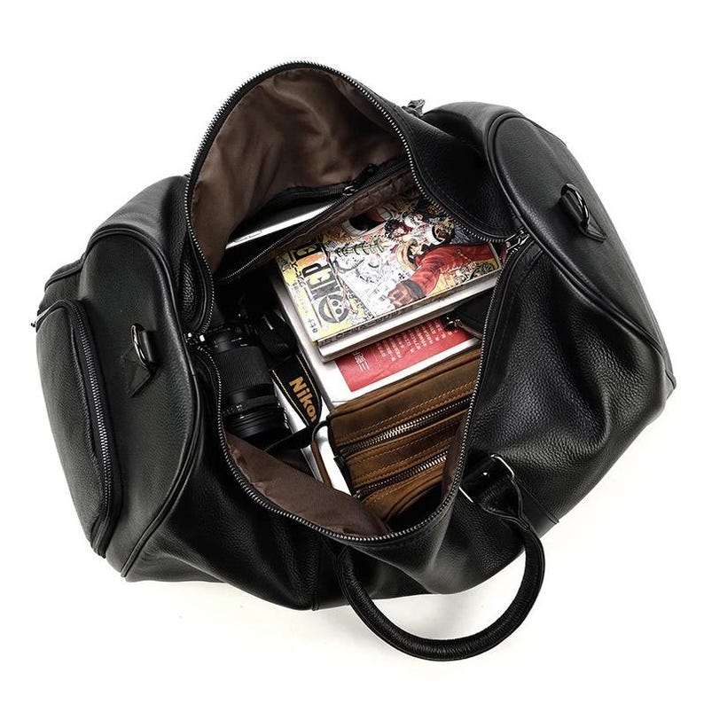 May include: A black leather duffel bag with a zipper closure and a top handle. The bag is open and filled with various items, including a camera, books, and a wallet.