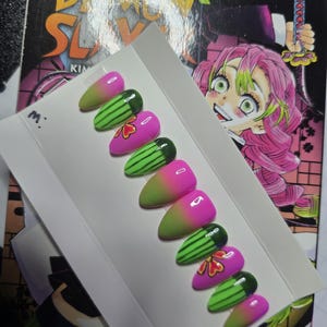 May include: A set of almond-shaped false nails. The nails feature a gradient design, transitioning from green to pink. Some nails are decorated with green watermelon patterns and red flower accents. The background includes a comic book.