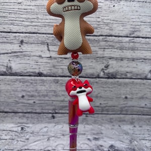 May include: A decorative pen featuring a brown and white fox-shaped topper with an expressive face. The pen has a metallic pink and purple barrel, and a smaller red fox charm. The pen is a unique and fun writing accessory.