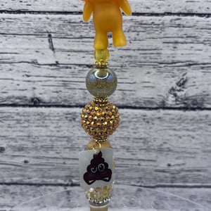 May include: A novelty pen with a yellow, smiling poop emoji figure at the top. The pen features decorative beads in gold and iridescent colors, and a small image of a poop emoji near the pen tip.