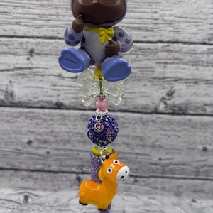 May include: A decorative pen featuring a baby figurine in a giraffe costume, a glittery ball, and an orange giraffe charm. The pen has a blue barrel and is topped with a clear, decorative element.