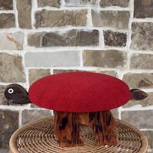 May include: A decorative turtle-shaped stool with a red cushion top. The turtle's head and flippers are made of dark wood, and the base is a carved wooden structure. The stool sits on a woven wicker base, with a brick wall in the background.