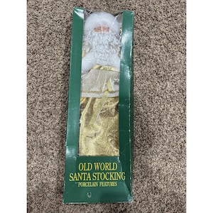 May include: A Santa Claus figurine with a white beard and hat, wearing a gold patterned garment, packaged in a green box labeled OLD WORLD SANTA STOCKING PORCELAIN FEATURES. The figurine has a porcelain face and is visible through a clear plastic window.