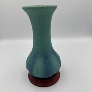 Puede incluir: Un jarrón de cerámica de color verde azulado con una base bulbosa y un cuello acampanado. La base tiene un diseño texturizado en forma de hoja. El jarrón se asienta sobre una base redonda de madera marrón oscuro, creando contraste.