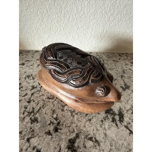 May include: A carved wooden sculpture in shades of brown. The top features a woven pattern, while the base has a smooth, rounded shape. The sculpture has a unique, organic form, possibly a decorative piece.