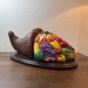 May include: A ceramic cornucopia sculpture featuring a brown woven-textured horn overflowing with a bounty of colorful fruits and vegetables, including a large orange pumpkin, purple grapes, yellow pears, and red apples, set on a dark oval base.